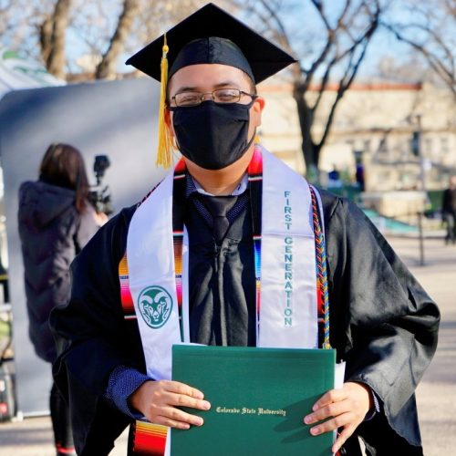 Masked student wearing graduation regalia and stole reading first generation