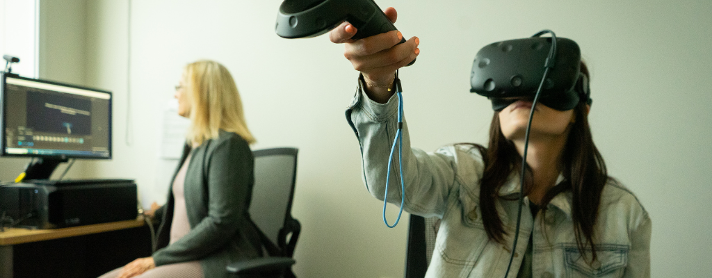 A psychology researcher monitors a computer in the background, as a student uses a VR headset in the foreground.