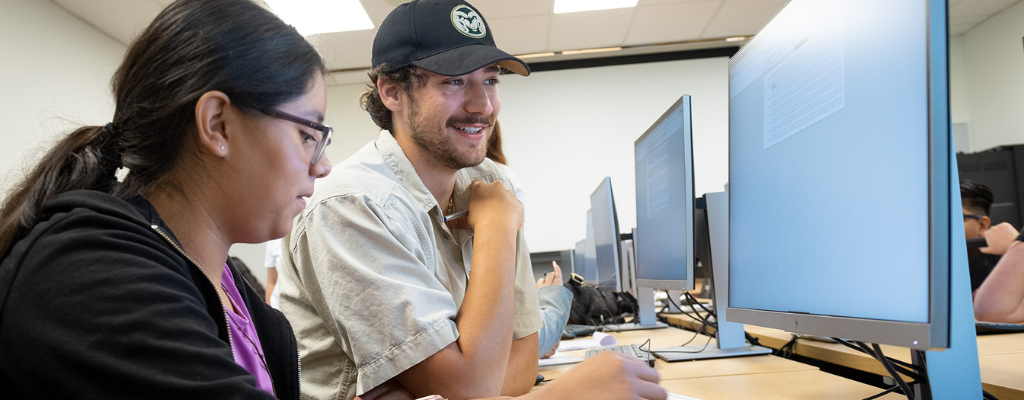 A CSU student in a CSU baseball cap helps a younger student during a Computer Science Summer Camp in 2022.