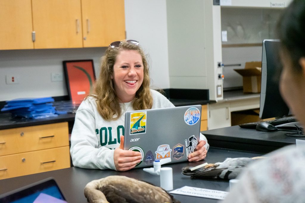Student smiles while working on a laptop and talking to another student.