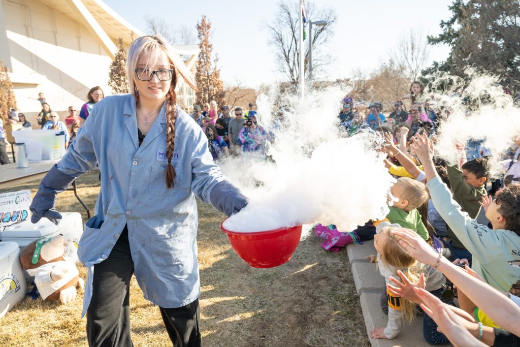 A student in a lab coat walks by a crowd of young students with a bowl of dry ice. The crowd reaches out to touch the fog.  
