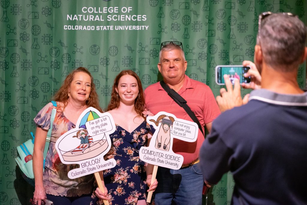 Someone takes an iPhone picture of a student posing with their parents holding two signs with cartoon rams that say "Biology" and "Computer Science."