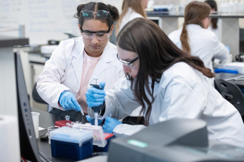Two students in lab coats pipette liquid into small vials.